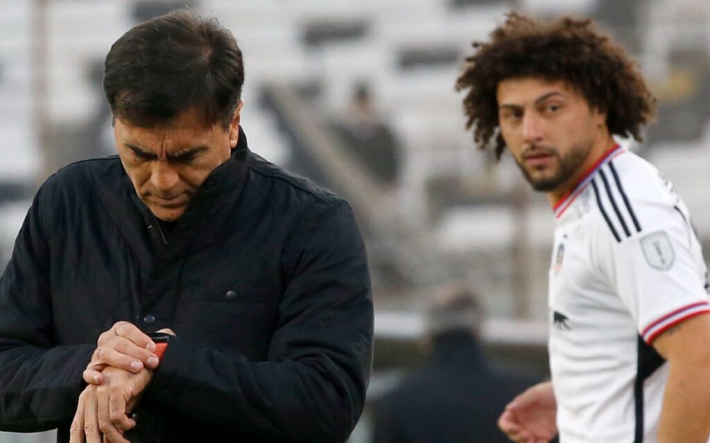 Gustavo Quinteros mirando su reloj y Maximiliano Falcón mirándolo en el fondo