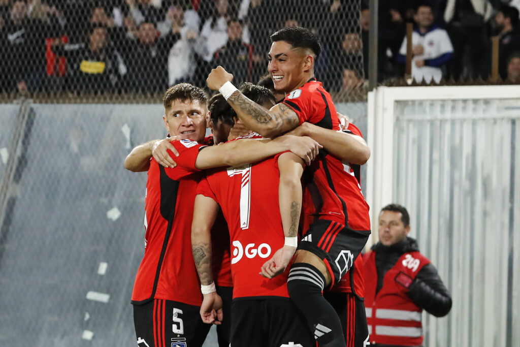 Plantel de Colo-Colo celebrando uno de los goles de Jordhy Thompson ante Unión la Calera, cuyo jugador es sostenido por un futbolista albo mientras empuña su mano en alto.