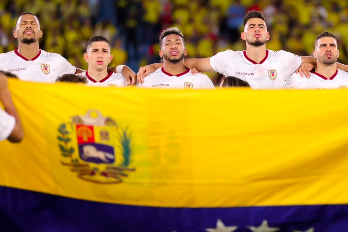 Futbolistas de la Selección de Venezuela se abrazan mientras entonan su himno nacional con la bandera de fondo.