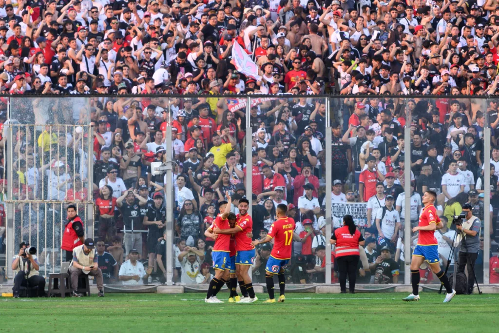 Futbolistas de Unión Española celebran un gol en la cancha principal del Estadio Monumental, mientras que en el fondo aparecen los hinchas de Colo-Colo en el sector Arica.