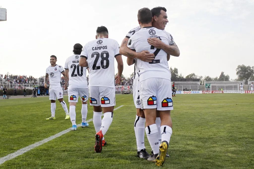 Esteban Paredes abraza a Gabriel Costa tras anotar un gol con la camiseta de Colo-Colo durante la temporada 2019 en el Estadio Municipal de La Cisterna, mientras que el resto de sus compañeros camina al círculo central.