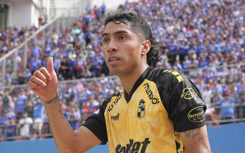 Luciano Cabral levanta su pulgar en pleno partido con l camiseta de Coquimbo Unido y la hinchada de Universidad de Chile de fondo.