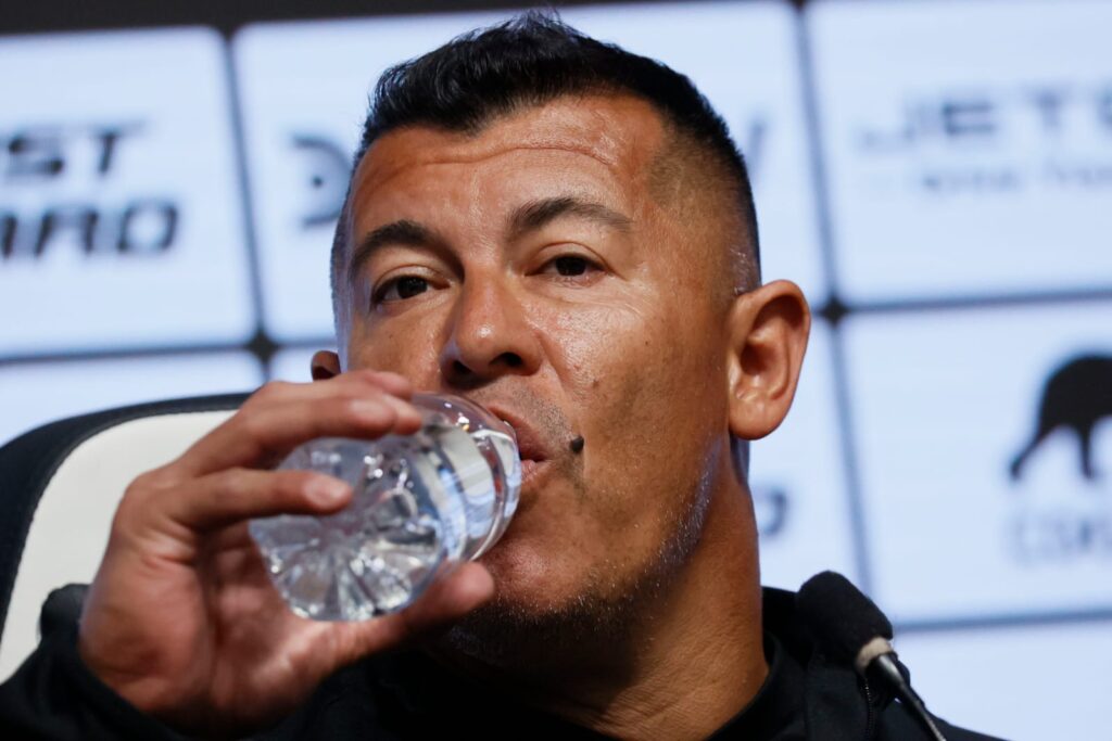 Primer plano a Jorge Almirón bebiendo agua desde una botella en una conferencia de prensa.